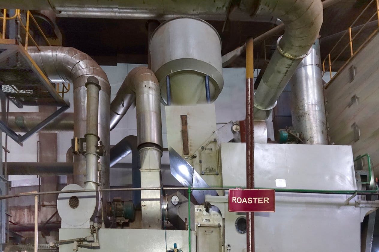 Industrial roaster on a continuous-process soluble coffee production line; large overhead ductwork dominates the upper portion of the frame; ROASTER equipment sign visible on the central machinery