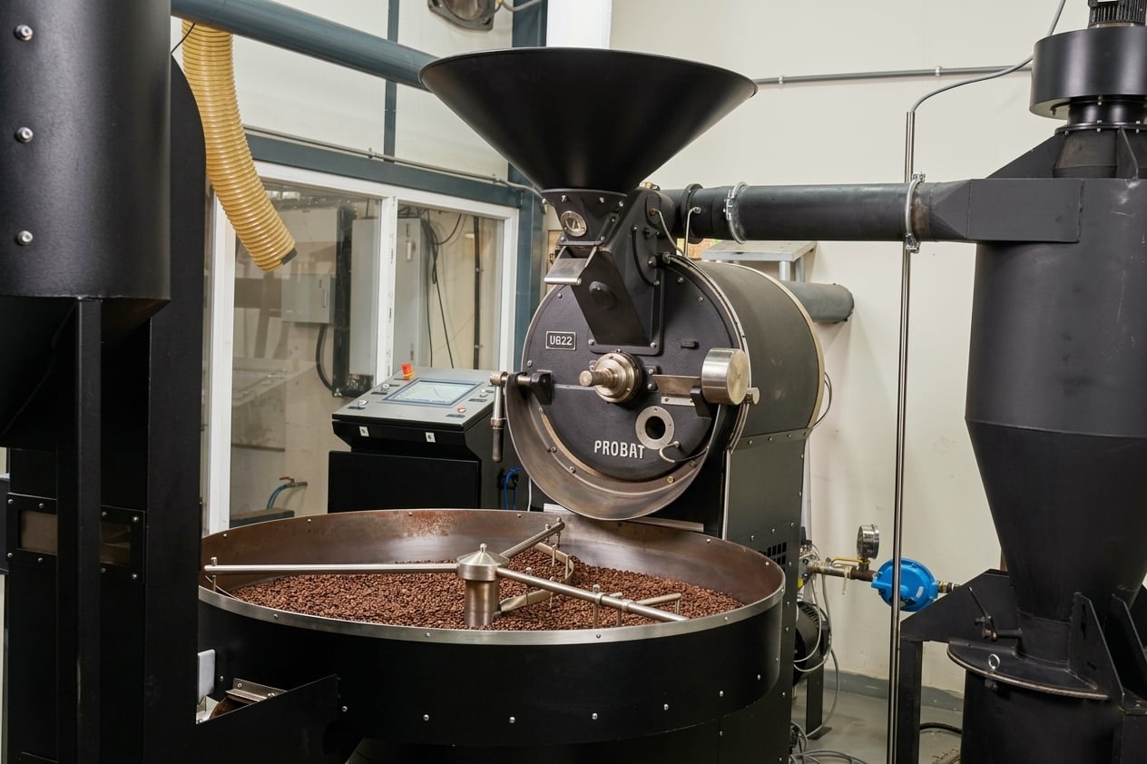 Specialty coffee roaster on a production floor — drum, hopper, control panel, and cooling tray of roasted beans visible.