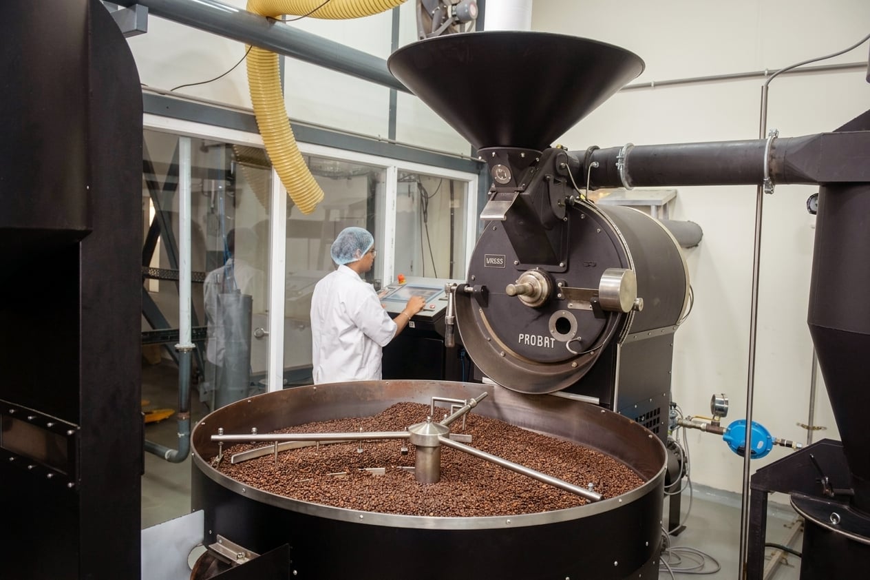 Specialty coffee roaster on the production floor; an operator in white lab coat works the control panel; cooling tray of roasted beans in the foreground